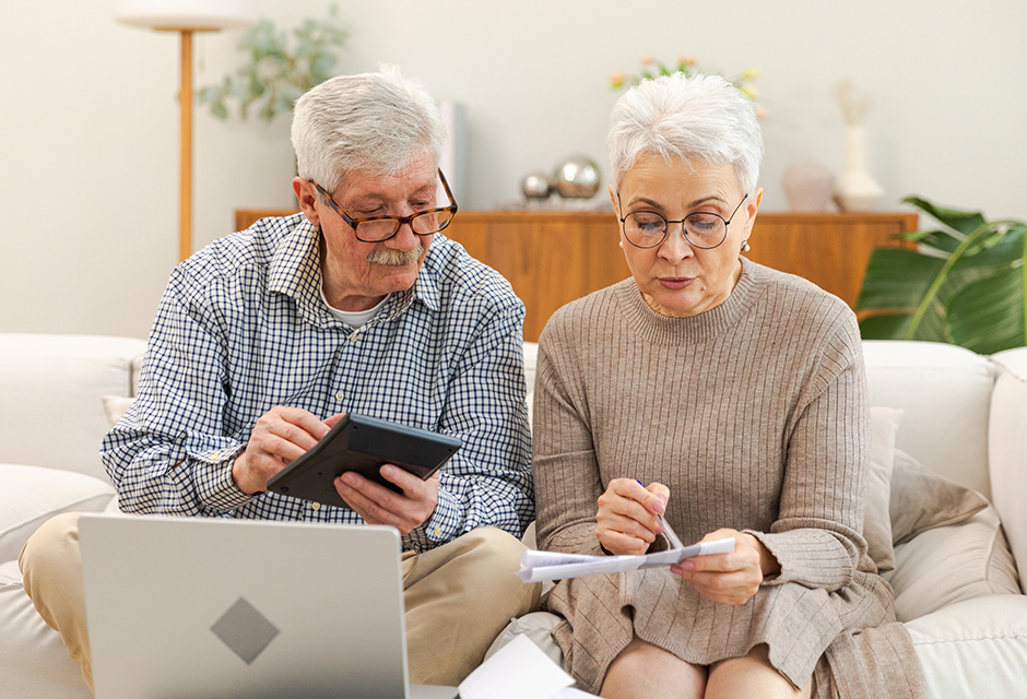 Older couple using a calculator and reviewing paperwork on a laptop while comparing Social Security lump-sum election calculations.