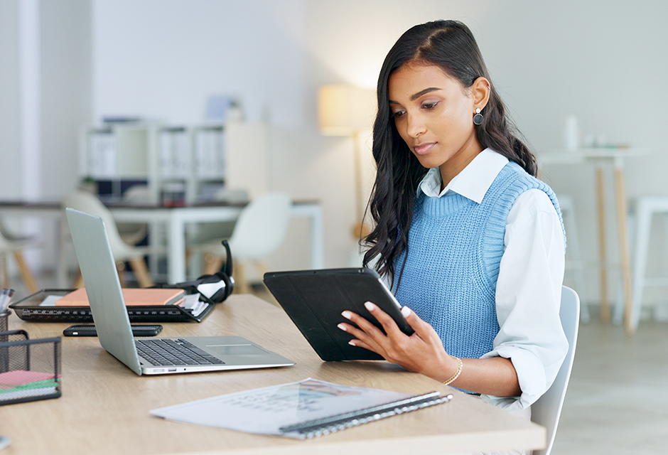 Woman reviews documents on a tablet beside a laptop, reflecting federal and state disaster loss analysis.