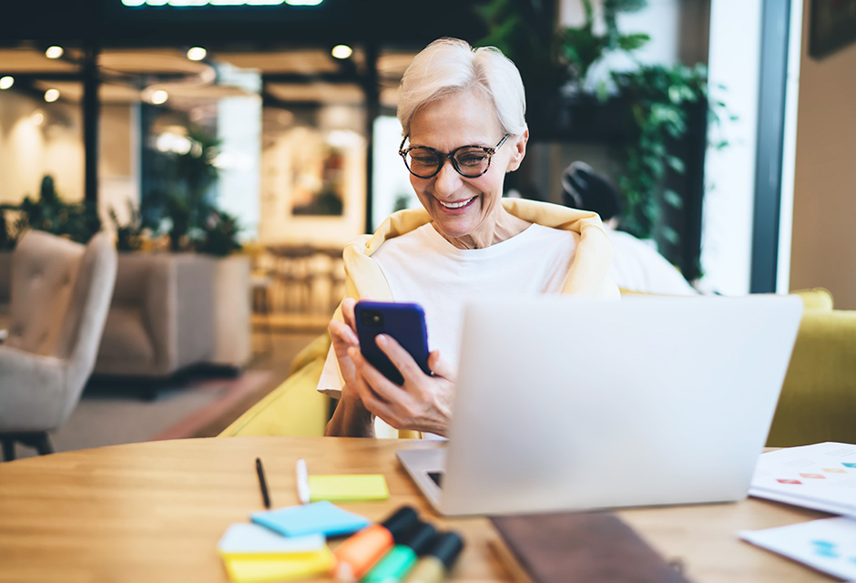 Older woman smiling at her smartphone beside a laptop, illustrating retirement tax planning and Roth conversion decisions.
