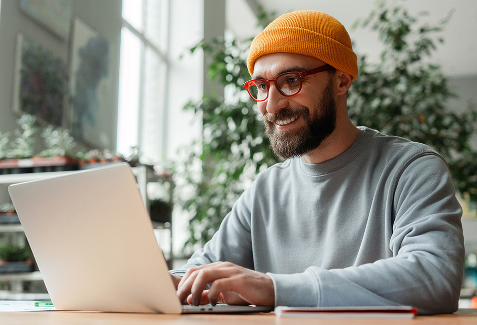 Man wearing a yellow hat and red glasses smiles while working on a laptop in a bright indoor space