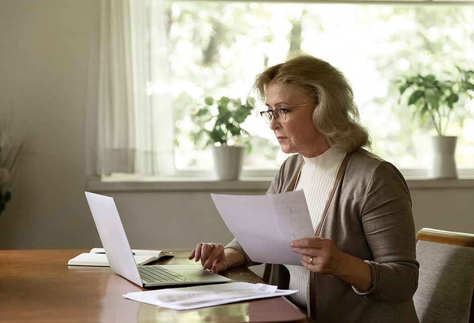 Older woman reviewing tax documents at a desk while working on a laptop, organizing paperwork for year-end tax planning.