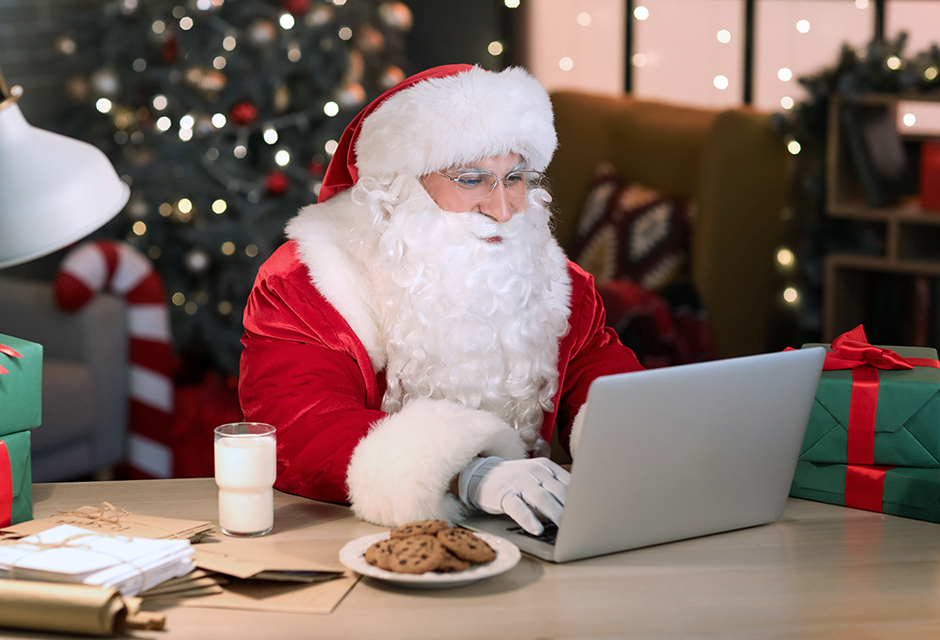 Santa Claus in a red suit working on a laptop at a desk with cookies and milk, symbolizing holiday tax planning and OBBBA-related tax questions for 2025.