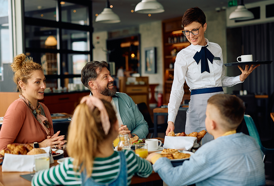 Smiling restaurant server delivering food to a family at their table, illustrating tipped work relevant to new 2025 qualified tip deductions.