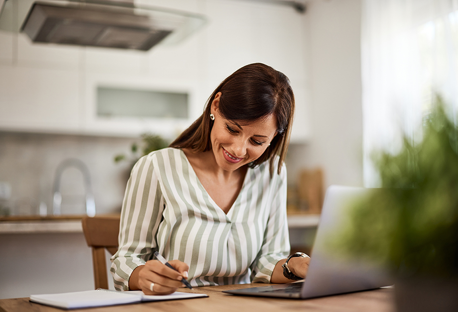 A woman sits at a kitchen table writing notes while working on a laptop, representing thoughtful charitable tax planning